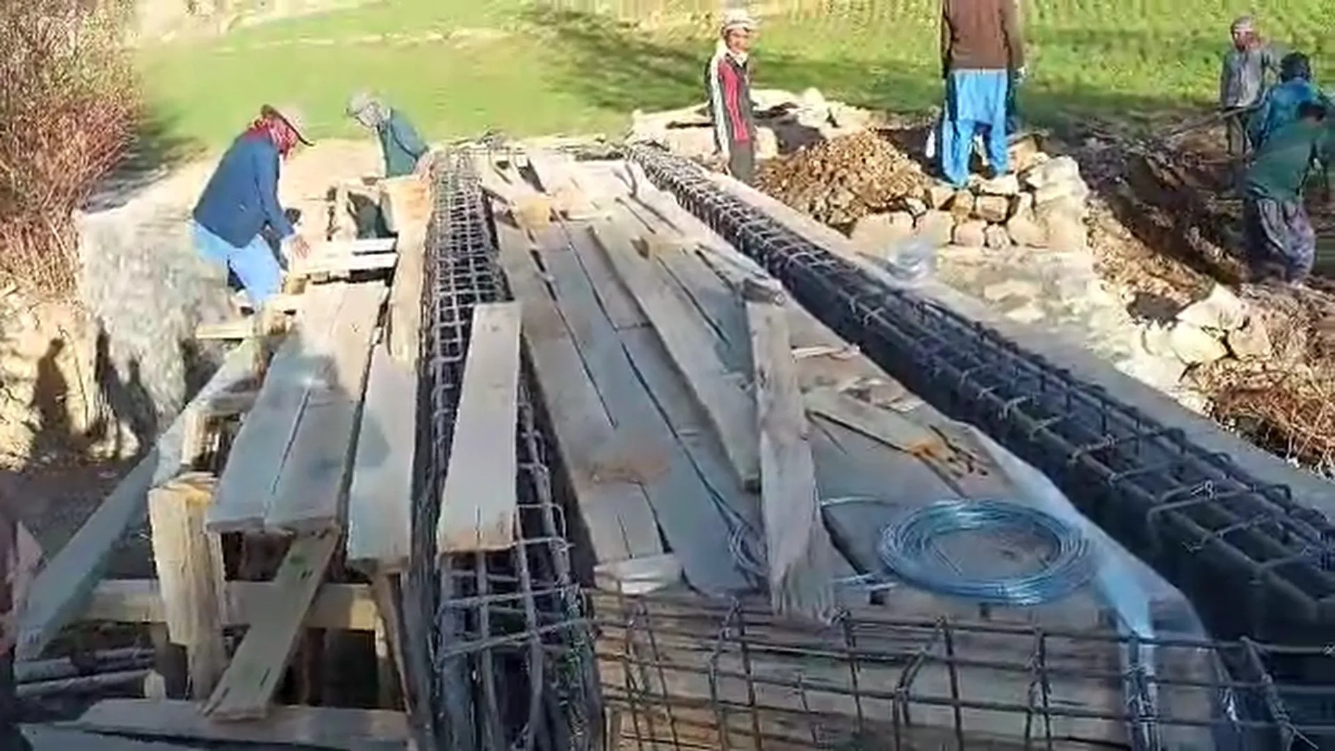 Arbeiter verlegen Armierungseisen und Holzbretter für die neue Brücke; daneben liegen Steine und Erde.