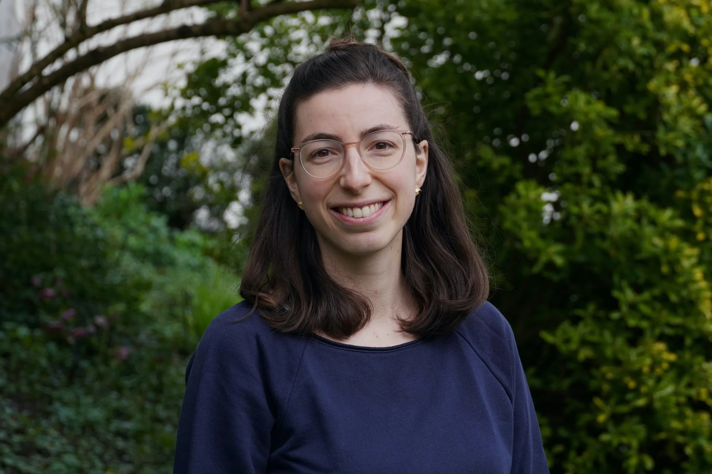 Nadine Lützelschwab mit Brille und dunkelblauem Oberteil lächelt vor grünen Sträuchern in die Kamera.