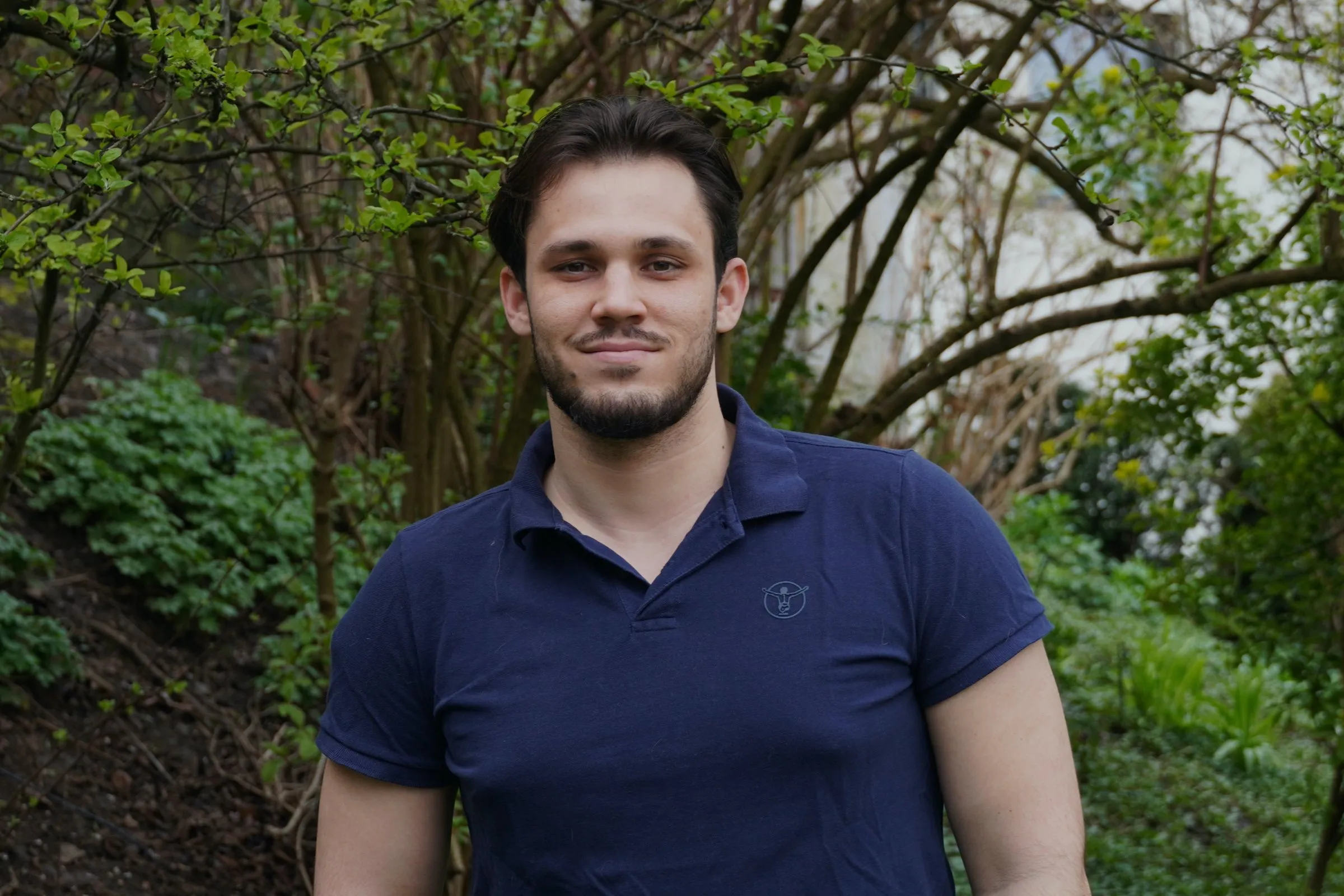 Noah Zilic im dunkelblauen Poloshirt lächelt vor Bäumen und Sträuchern in die Kamera.