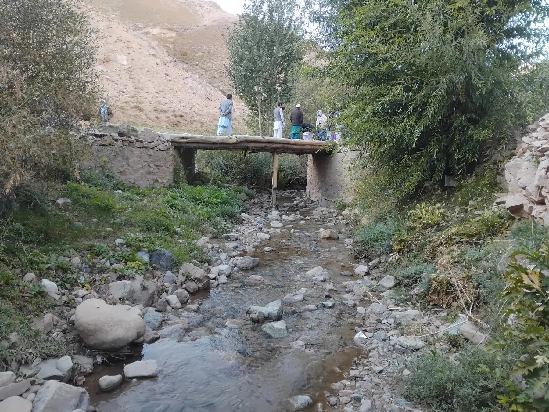 Die alte Brücke – ein schmaler Übergang aus Holzbalken auf zwei Steinmauern über einem steinigen Bach; darauf stehen einige Männer.