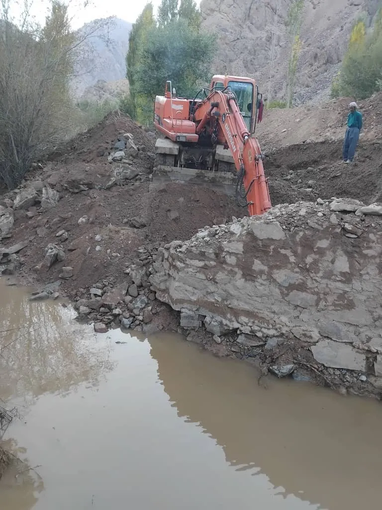 Ein orangefarbener Bagger hebt neben einem Mauerrest Erde aus dem Flussbett; im Vordergrund steht braunes Wasser.