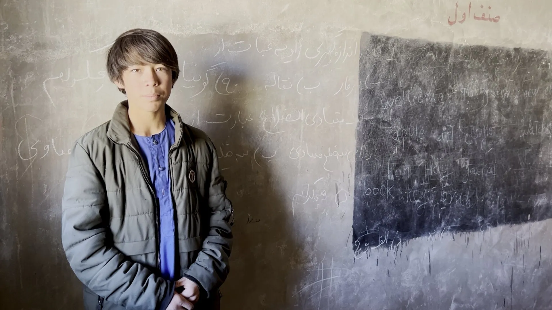 Ein Schüler steht vor einer Wand, die in mehreren Schichten mit persischer und englischer Schrift bedeckt ist; daneben eine aufgemalte schwarze Tafel.