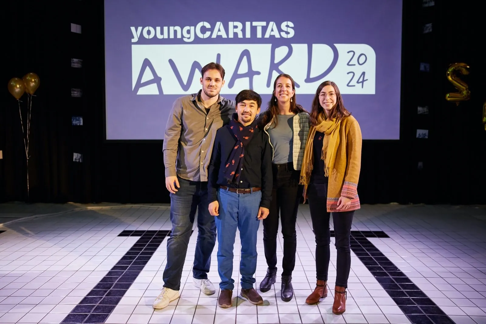 Vier Mitglieder von Compassion in the Right Place stehen auf der Bühne vor der Leinwand mit dem Schriftzug «youngCARITAS Award 2024».
